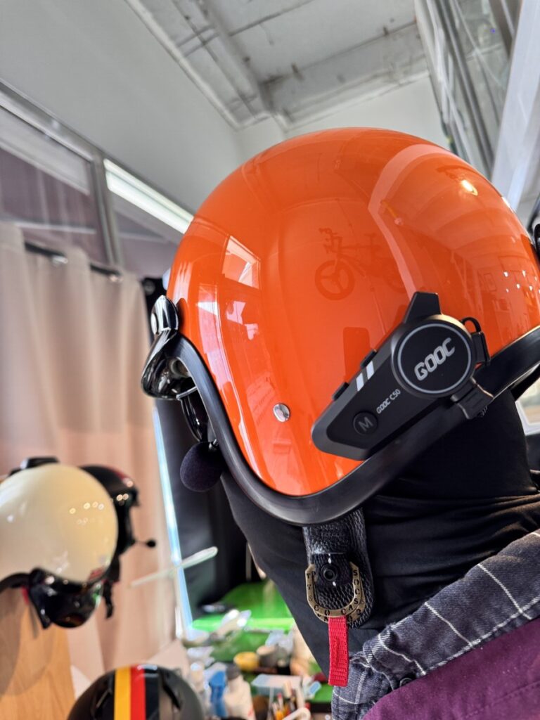 "Person wearing a bright orange open-face motorcycle helmet equipped with a GOOC C50 Bluetooth intercom, viewed indoors with other helmets in the background."