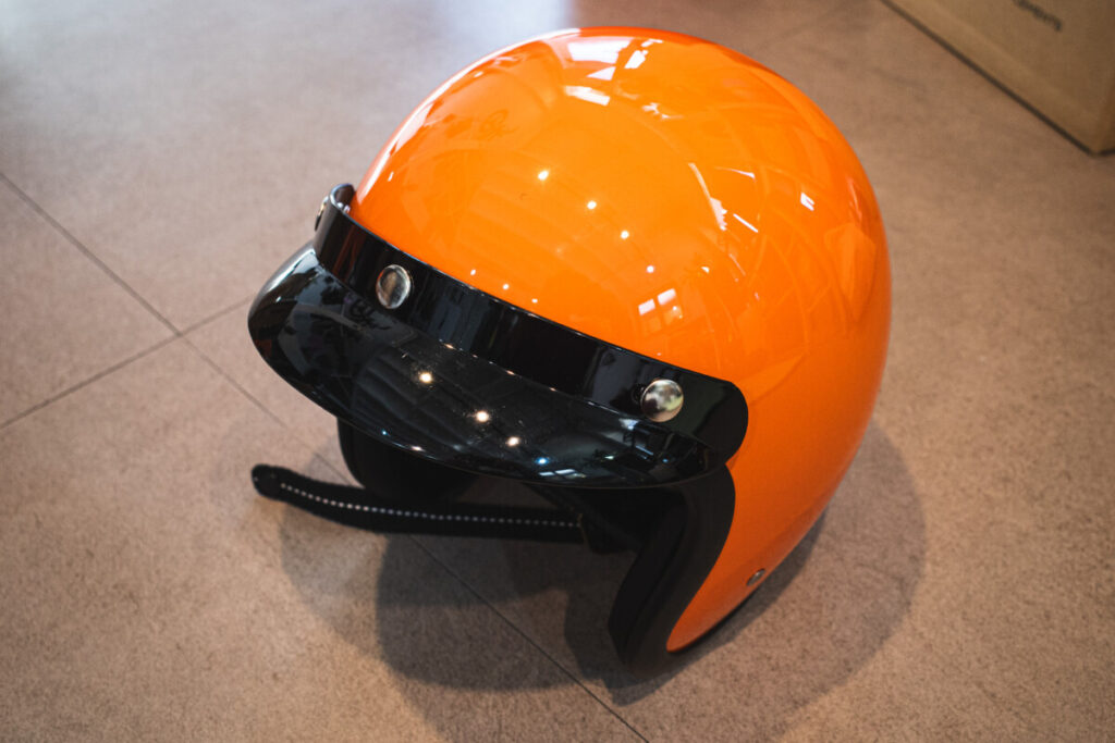 "Glossy orange open-face motorcycle helmet with a black visor and chin strap, placed on a tiled indoor floor."