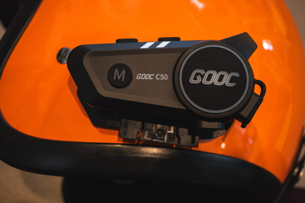 "GOOC C50 Bluetooth intercom mounted on an orange motorcycle helmet, showing the main control button and clip-on attachment clearly." 2
