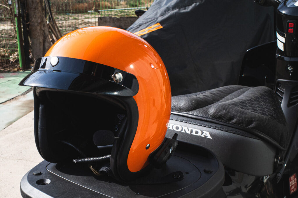 "Bright orange open-face motorcycle helmet with a black visor, placed on a Honda scooter seat outdoors in daylight."