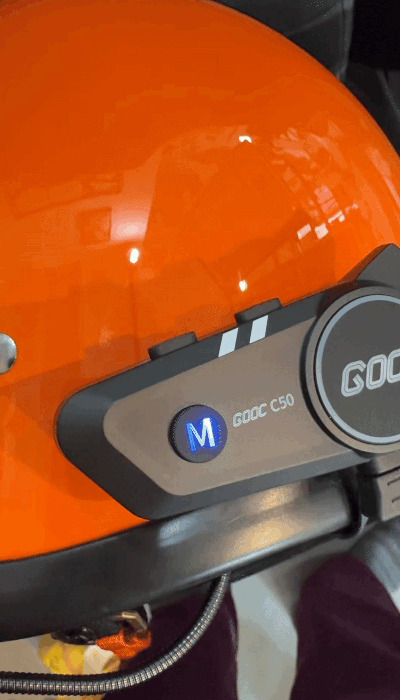 "Animated view of the GOOC C50 Bluetooth intercom mounted on an orange motorcycle helmet, showing the illuminated red 'M' button during power-on or pairing mode."