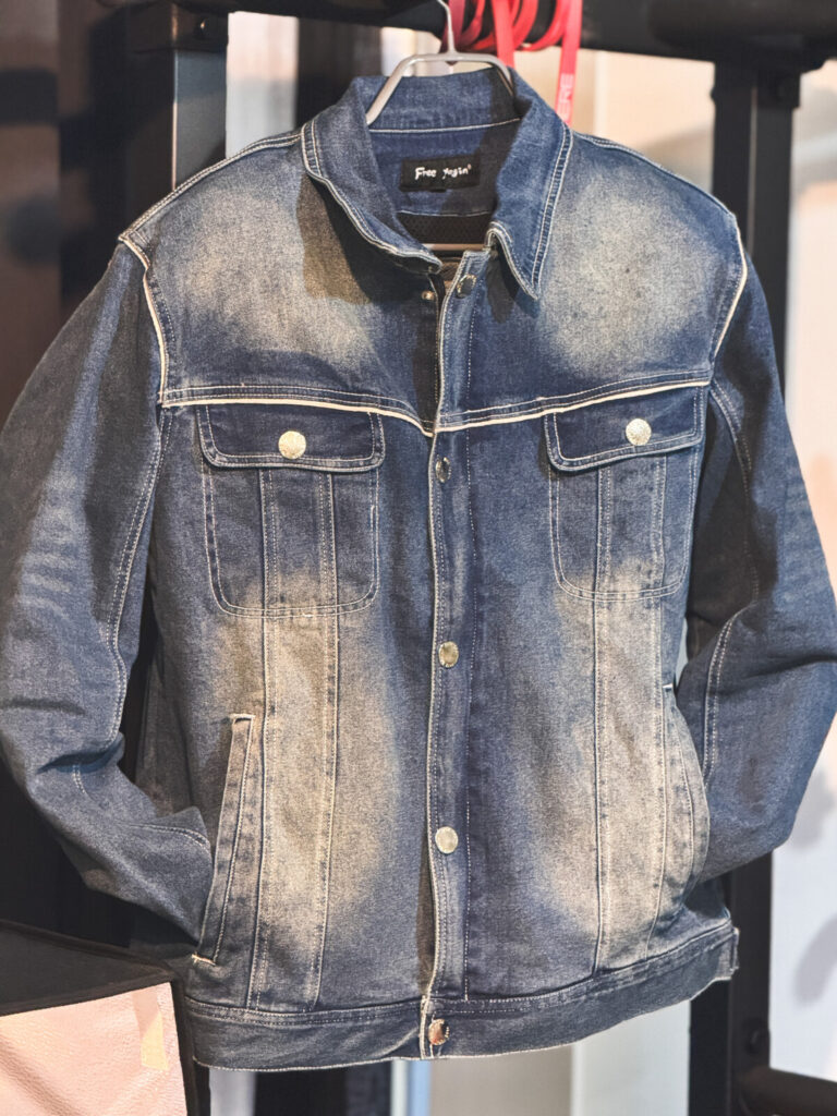 "Close-up front view of a denim jacket hanging on a hanger, showing chest flap pockets, faded wash, and 'Free Again' label on the collar"