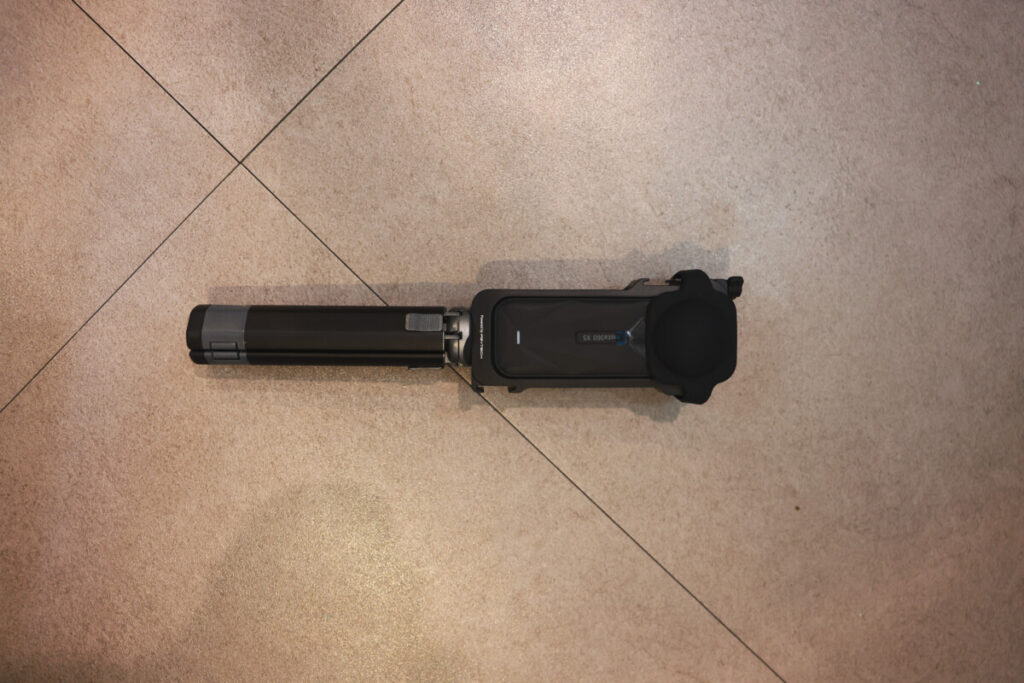 “Top-down view of the Insta360 X5 camera with its lens cap on, attached to a folded selfie stick, placed on a tiled floor.”
